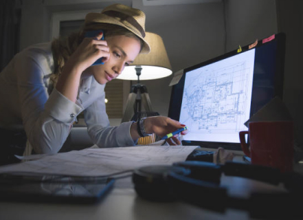 A women in a fedora, talks on the phone while comparing paper drawings to ones on her screen.